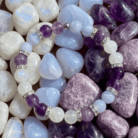 Pulsera Serena - Alivio Natural para la Menopausia, con cuentas moradas, blancas y translúcidas, separadores plateados y exhibida sobre piedras en tonos púrpura, azul y blanco para aliviar los síntomas de la menopausia.